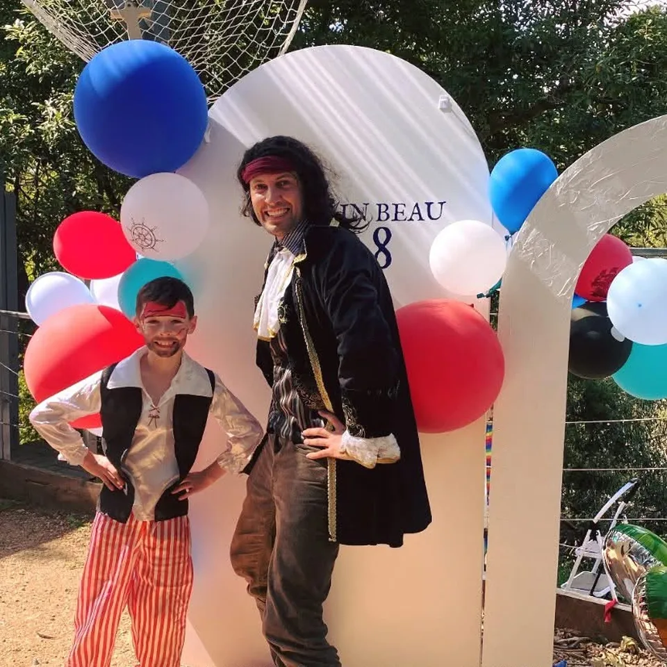 Pirate party entertainer in full captain costume with bandana and ruffled shirt posing with young boy in pirate vest, striped pants and pirate face paint beside red, white and blue balloon arch at a Melbourne pirate themed birthday party