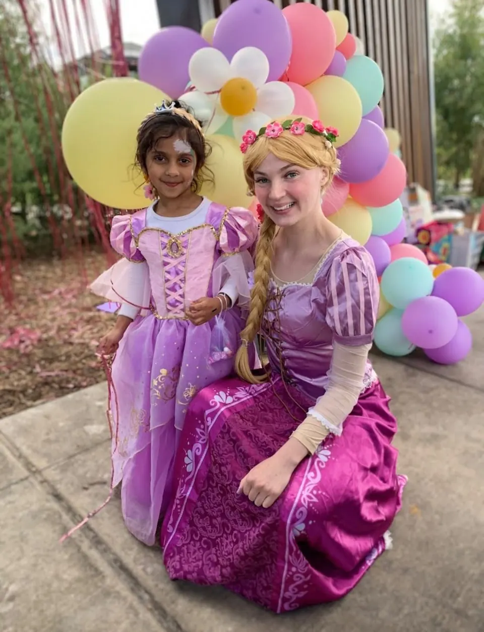 Princess Rapunzel party entertainer with long blonde braid and purple Tangled dress posing with young girl in Rapunzel costume beside pastel balloon garland in Melbourne