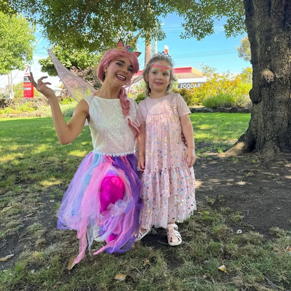 Unicorn fairy party entertainer with pink hair, gold horn and fairy wings posing in a sunny park with young girl in tiara, sparkly pink dress and rainbow unicorn face paint at a Melbourne outdoor unicorn fairy birthday party