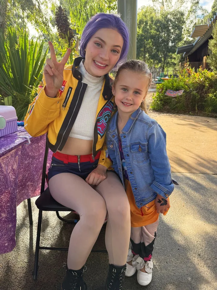 K-Pop Demon Hunters Rumi party entertainer in purple wig and yellow Huntr/x jacket cuddling young girl beside a purple party table at a Melbourne outdoor K-Pop themed birthday party