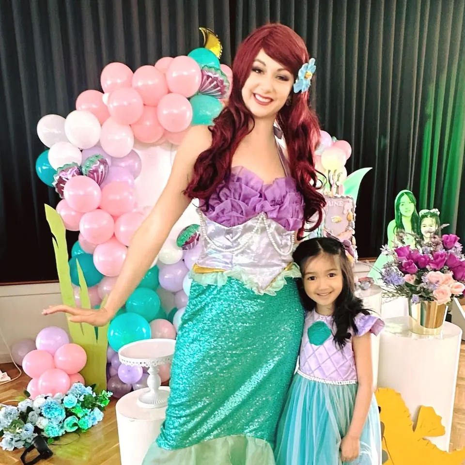 Little Mermaid Ariel party entertainer in green sequin tail with young girl in mermaid dress beside pink and teal balloon arch in Melbourne