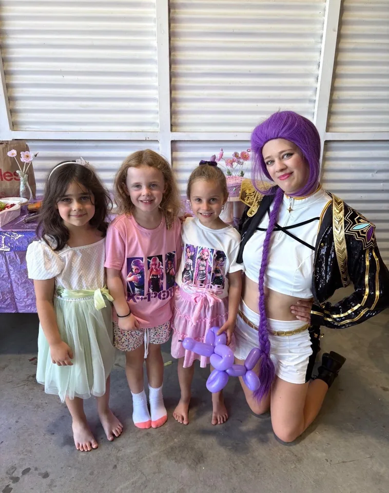 K-Pop Demon Hunters Mira party entertainer with long purple braid posing with three young girls wearing K-Pop Demon Hunters Huntr/x T-shirts at a Melbourne K-Pop birthday party